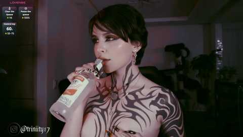 Video of a woman with short dark hair, heavily tattooed chest, drinking from a water bottle, in a dimly lit room with plants, social media app screen overlay.
