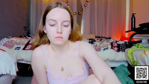 Video of a young white woman with long brown hair, wearing a light pink lace bralette, sitting on a bed with a patterned quilt and fairy lights in the background.