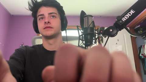 Video of a young man with short black hair, wearing headphones, recording with a RODE microphone in a purple room.