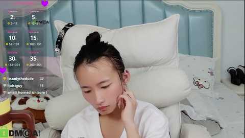 Video of a young Asian girl with dark hair tied up, wearing a white robe, sitting on a white bed with a light blue padded headboard. She's eating chocolate and looking at her phone.