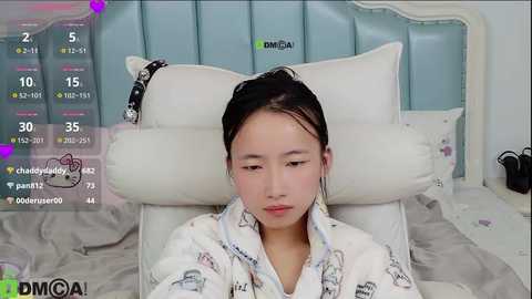 Video of an East Asian woman with straight black hair, wearing a white bathrobe with cartoon characters, lying on a white bed with a light blue headboard. Digital overlay shows heart icons and statistics.
