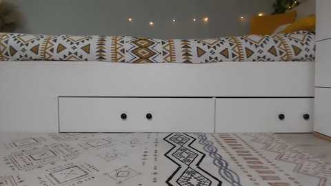 Video of a modern, minimalist bedroom featuring a white bed with geometric patterned bedding and a matching white dresser with two drawers, set against a soft grey wall.