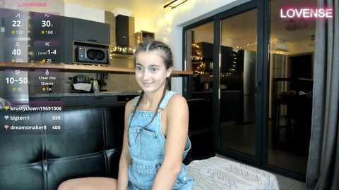Video of a young woman with light skin and dark hair in overalls, smiling in a modern kitchen with a black couch.