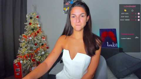 Video of a young woman with straight, dark hair in a strapless white dress, sitting in a modern living room with a decorated Christmas tree, a large screen TV, and a plush sofa.