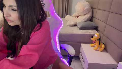 Video of a woman with long brown hair in a pink jacket, standing beside a white chair. A plush yellow dog toy sits on a white nightstand in a modern, beige-tiled bedroom.