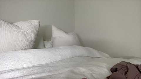 A video of a minimalist, white bed with a textured quilt, two white pillows, and a crumpled brown garment on the floor. The walls are plain, light gray, creating a serene, uncluttered atmosphere.