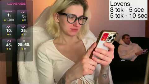 Video of a blonde woman with glasses in a white dress, holding a phone, watching a man in a white shirt recline on a couch in the background.