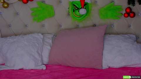 A video of a bed with a plush green, grinning mask of Grinch and colorful ornaments hanging above, surrounded by white and pink pillows.