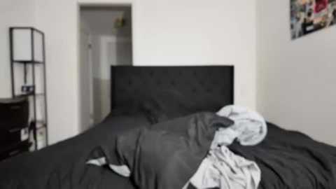 A video shows a messy bedroom with a black upholstered headboard, disheveled gray bedding, a black shelf, and a partially open doorway.