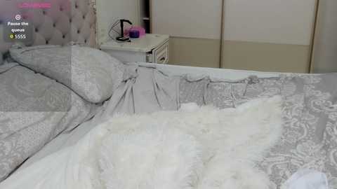 Video of a plush, tufted, white bed with grey bedding, fluffy white throw, and a small white nightstand with a fan and lamp in a minimalist, modern bedroom.