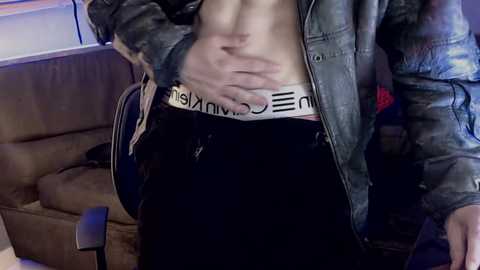 Video of a shirtless man in black Calvin Klein underwear, denim jacket, and black pants, sitting on a brown couch in a dimly lit room.