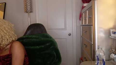 A video of a woman with a curly blonde wig and dark hair, wearing a green robe, standing in a cluttered bathroom with a white door, a mirror, and a cluttered vanity.