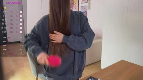 Video of a woman with long, straight brown hair in a dark blue sweater, brushing her hair with a red brush in a modern living room.