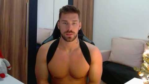 Video of a muscular, shirtless man with short brown hair, beard, and black harness, seated in a gaming chair, in a modern room with beige cushions and wooden cabinets.