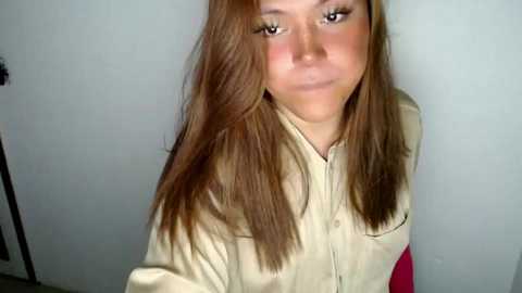 Video of a young woman with fair skin and straight, light brown hair, wearing a beige button-up shirt, standing against a plain, light gray wall.