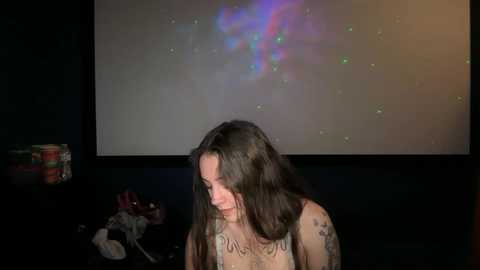 Media: Video of a woman with long brown hair, wearing a sleeveless top, sitting in a dimly lit room. Behind her, a large TV screen displays a colorful, abstract light show.
