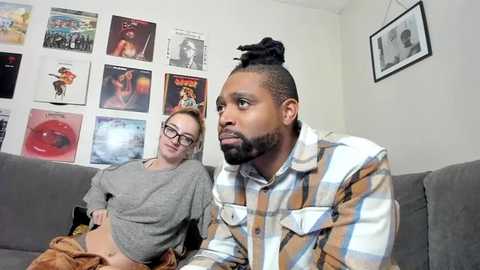 Media: Video of a young man with dreadlocks and a woman with glasses, both sitting on a gray couch, watching a TV in a room with a wall of posters and framed photos.
