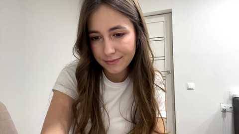 Video of a young woman with long brown hair, wearing a white t-shirt, smiling softly in a white-walled room with a door and light switch visible in the background.