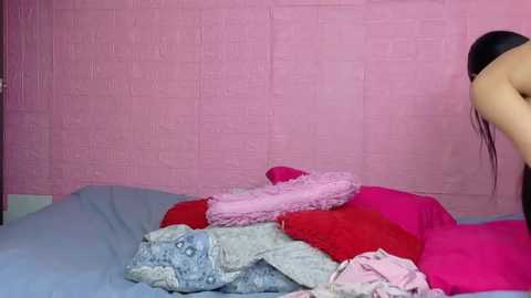 Video of a young woman with long black hair, partially visible, standing in a pink brick-walled room, wearing a pink bra and panties. She's arranging clothes on a bed with blue sheets and pink and red blankets.
