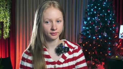 A video of a young girl with straight blonde hair, wearing a red and white striped sweater, standing in front of a Christmas tree with colorful lights and a potted plant in the background.