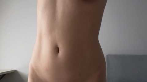 A video of a nude, fair-skinned torso with a flat abdomen, visible navel, and no visible genitalia. The background features a light gray wall and a corner of a white object, likely a piece of furniture.