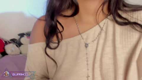 Video of a woman with medium brown skin, long dark hair, wearing a beige off-shoulder sweater, a silver necklace with a cross, and a rosary. Background features a stuffed panda and a bed with a purple pillow.