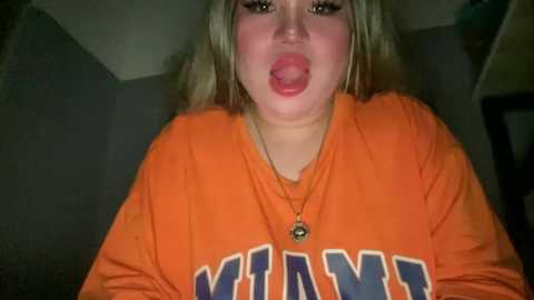Video of a young woman with fair skin and blonde hair, wearing an orange University of Miami sweatshirt, sticking her tongue out, indoors with dim lighting.