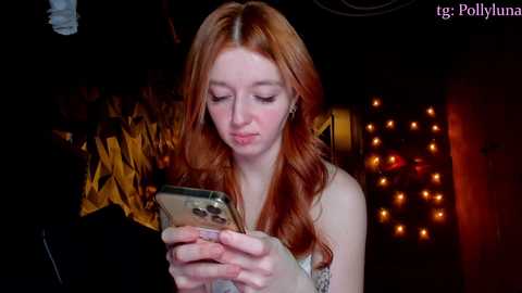 Media: Video of a young woman with long red hair, wearing a sleeveless top, intently looking at her phone in a dimly lit room with warm, glowing string lights in the background.
