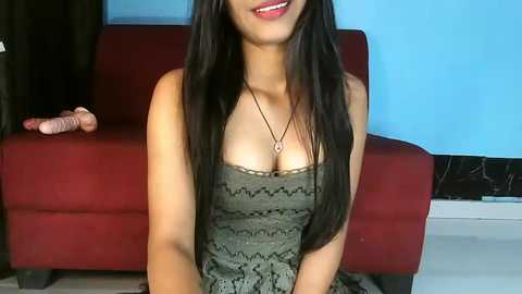 Media: Video of a young Asian woman with long black hair, wearing a low-cut green lace dress, sitting on a red couch, smiling, against a blue wall.