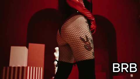 Video of a woman with long black hair, wearing red latex gloves and fishnet stockings, standing against a red backdrop. Her buttocks are prominently exposed, with the fishnet revealing her skin.
