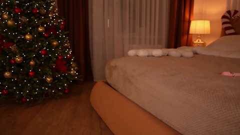 Video of a warmly-lit bedroom with a decorated Christmas tree on the left, a neatly made bed with white pillows and a beige bedspread on the right, and a cozy lamp beside it.