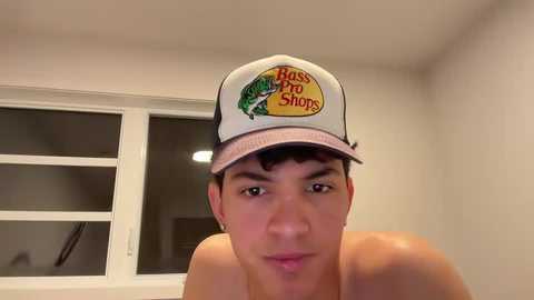 Video of a shirtless young man with medium build, light skin, and short dark hair, wearing a \"Boise Diner\" cap indoors. Background includes a white-framed window with darkened glass and beige walls.