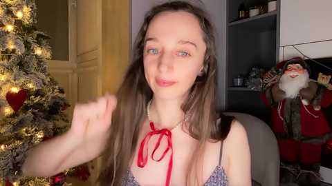 A video of a young woman with long brown hair, fair skin, and blue eyes, wearing a red ribbon necklace, playfully touching her hair. She stands next to a decorated Christmas tree in a cozy, warmly lit room.