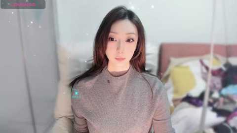 Video of an East Asian woman with straight black hair, wearing a gray long-sleeve top, standing in a bedroom with a white bed and colorful pillows.