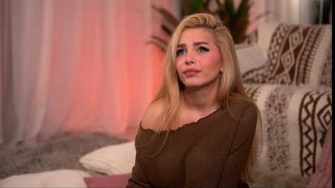 Video of a blonde Caucasian woman with long hair, wearing a brown off-shoulder top, looking thoughtful, in a cozy room with patterned cushions, pink and white curtains, and a green plant.