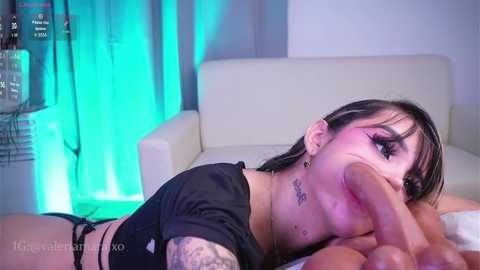Video of a tattooed woman with black hair, wearing a black crop top, performing oral sex on a large, erect penis in a dimly lit room with blue lighting.