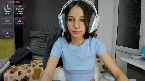 Video of a young woman with light skin and long brown hair, wearing a light blue shirt, white headphones, sitting in a messy bedroom.