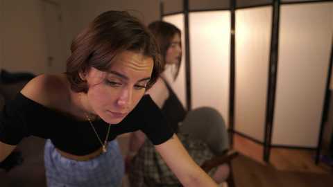 Video of a woman with short, dark hair, wearing a black top and light blue apron, bending over a wooden table in a dimly lit room with a sliding paper screen in the background.