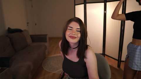 Video of a smiling young woman with medium-length brown hair in a black tank top, seated in a living room with a beige sofa and wooden floor.