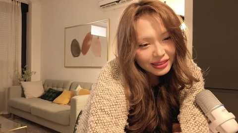 Media: Video of an Asian woman with long brown hair, wearing a beige knit cardigan, smiling while holding a microphone in a modern living room with a gray couch, abstract art, and white walls.