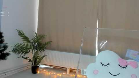 Video of a cozy room with a potted fern, a radiator, and a clear glass table with a plush cloud pillow featuring a smiling face and pink bow.