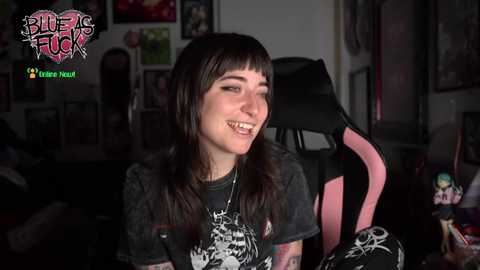Media: Video of a smiling, pale-skinned, tattooed woman with long dark hair and bangs, wearing a graphic t-shirt, sitting in a pink gaming chair in a dimly-lit room filled with posters and figurines.