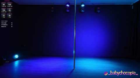 Video of a dimly lit, empty dance studio with a single pole in the center under blue and purple lights. Text overlay on the left includes \"Babychocolate\" logo.