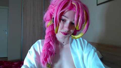 A video of a light-skinned woman with long, pink and yellow wig, wearing a white robe, standing indoors near a door and bed.