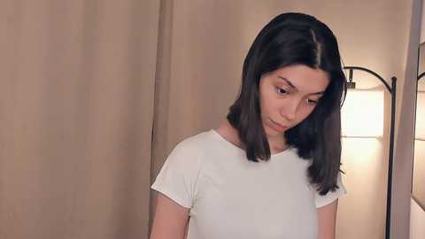Video of a young woman with long, straight black hair, wearing a white T-shirt, standing in a softly lit room with beige curtains and a modern floor lamp.