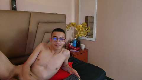 Video of a young, shirtless Asian man with glasses, lying on a bed with a brown headboard, in a bedroom with a mirror, yellow flowers, and a red pillow.