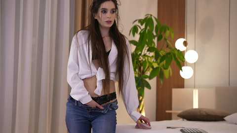 Video of a slender, brunette woman in a white tie-front blouse, black bra, and blue jeans, standing in a modern bedroom with a green plant, round lamp, and beige curtains.