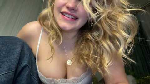 Video of a smiling, fair-skinned woman with wavy blonde hair, wearing a white lace bra and a necklace, leaning forward in a casual indoor setting.