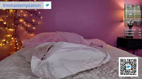 Video of a cozy bedroom with a white duvet, pink walls, string lights, and a digital clock on the nightstand. QR code overlay suggests augmented reality.