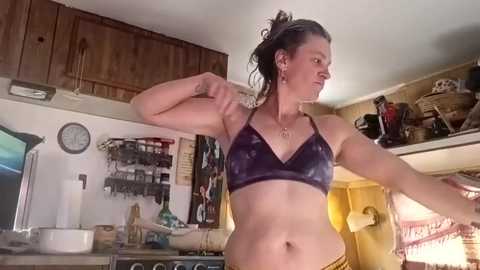 Video of a woman with a muscular build, wearing a tie-dye sports bra and yellow shorts, flexing her arm in a rustic, cluttered RV kitchen.